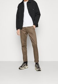 Veste noire légère, t-shirt blanc, pantalon slim marron et baskets noires et grises avec semelles texturées. Design simple et couleurs neutres.