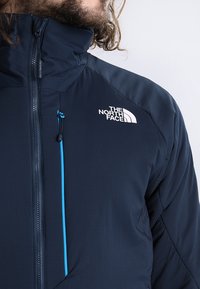 Marinblå jacka i texturerad tyg med full dragkedja, hög krage och en kontrasterande blå detalj på dragkedjan. The North Face-logotypen är synlig.