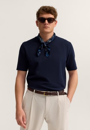 Mann mit dunkler Sonnenbrille, dunkelblauem Kurzarmhemd, dunkelblau gemustertem Halstuch, beigen Hosen und braun geflochtenem Gürtel, steht mit einer Hand in der Tasche.