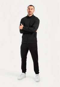 Zwarte jas met ritssluiting en hoge kraag, gecombineerd met zwarte broek. Model draagt witte sneakers. Soepele textuur, minimalistisch ontwerp.