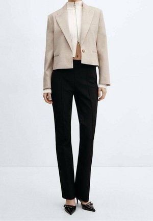 Blazer beige court porté sur un haut blanc à fermeture éclair, pantalon noir slim et escarpins noirs à bout pointu sur une personne debout devant un fond blanc.