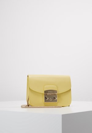 Lille gul læder crossbody taske med guldlås og kæderem, placeret på en hvid geometrisk piedestal mod en ensfarvet baggrund.