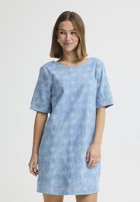 Femme aux cheveux mi-longs portant une robe bleu clair avec une broderie florale blanche, debout devant un fond uni clair.