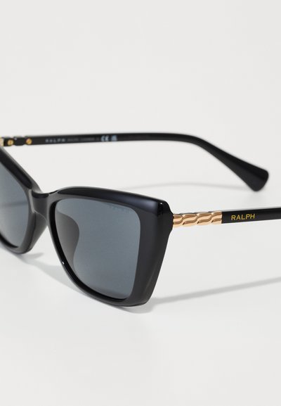 Gafas de sol negro estilo cat-eye con lentes oscuros; acentos dorados en las patillas; detalles texturizados cerca de las bisagras; nombre de la marca "RALPH" en amarillo.