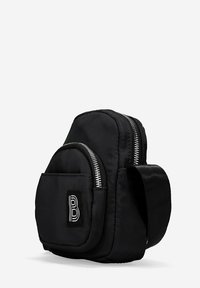 Mochila negra de nailon con diseño acolchado, cremalleras plateadas, bolsillo frontal y correa ajustable. Presenta un parche con logo en la parte delantera. Forma compacta.