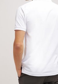 Homme portant un polo blanc uni à manches courtes, une main dans la poche d’un pantalon foncé, tourné de dos sur un fond neutre.