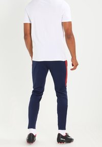Hombre con camiseta blanca, pantalones deportivos azul marino con raya roja, calcetines blancos y botas de fútbol negras, de pie contra un fondo blanco.