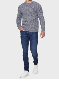 Šedý melírovaný svetr s kulatým výstřihem a žlutým emblémem, zkombinovaný s tmavě modrými upnutými džínami a bílými teniskami. Jednoduchý, strukturovaný vzhled.