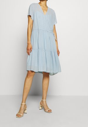 Femme portant une robe bleu clair mi-longue aux genoux avec manches courtes et des sandales blanches à talons hauts, debout devant un fond uni.