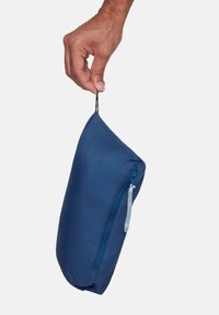 Mano che tiene una pochette compatta blu navy con zip tramite un piccolo anello di stoffa, su uno sfondo semplice.
