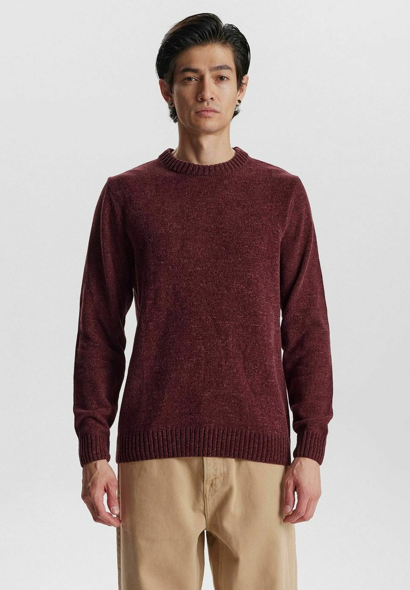 Burgunderfarbener Strickpullover mit geripptem Rundhalsausschnitt, langen Ärmeln und geripptem Saum. Der Stoff zeigt eine dezente Textur. Kombiniert mit beigen Hosen.