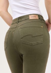 Olivově zelené džíny s hladkou texturou, designem s pěti kapsami a koženou nášivkou na pase s názvem značky "ANGELS."