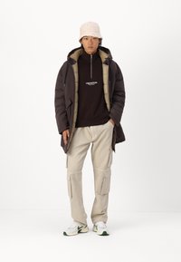 Abrigo acolchado marrón con forro beige, cuello con cremallera, sudadera negra con logo, pantalones cargo beige claros y zapatillas blancas con acentos verdes.