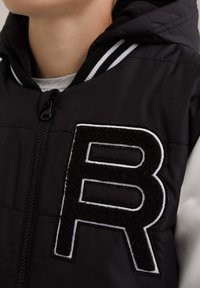 Jachetă neagră căptușită cu glugă, având un patch mare din fetru alb-negru cu litera "B" pe piept și manșete cu dungi ribbed.