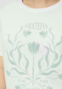 Witte katoenen T-shirt met een groen bloemenpatroon en de tekst "DANKBAAR VOOR DE MOOIHEID." Rond de hals en korte mouwen.