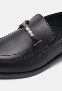 Mocassins en cuir noir au design épuré, avec un accent métallique portant le logo "BOSS", des détails cousus et une semelle texturée.