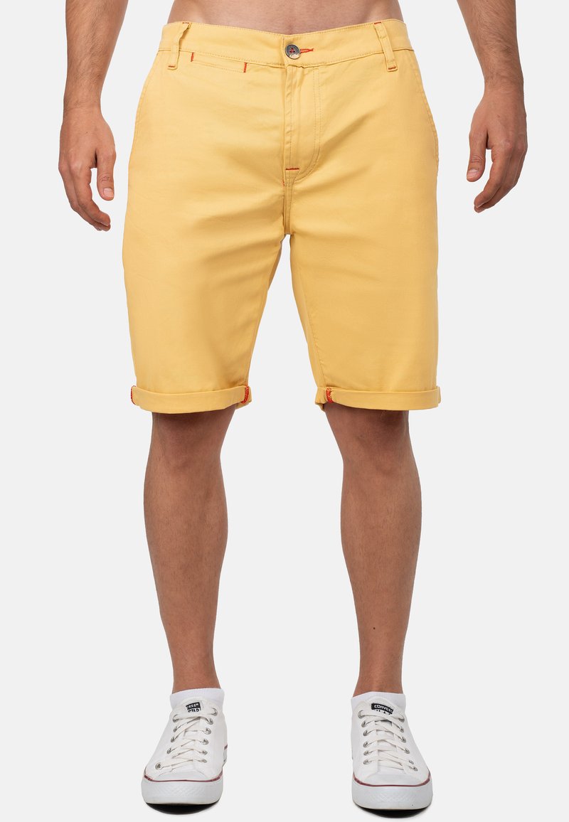 Gele katoenen shorts met een opgerolde zoom, met oranje stikdetails en een frontale knoopsluiting. Geshockeerd met witte sneakers.