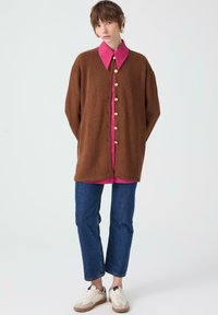 Brown ribbed oversized cardigan with large buttons, paired with a pink collared shirt, blue straight-leg jeans, and beige sneakers.