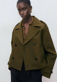 Woman wearing an oversized olive green double-breasted jacket with black buttons and gold hoop earrings against a plain background.