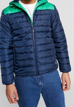 Homme portant une veste matelassée bleu marine avec des panneaux verts aux épaules, par-dessus une chemise blanche et un jean bleu, debout devant un fond uni.