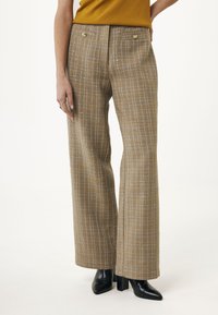 Femme portant un pantalon large à taille haute à carreaux avec des boutons dorés associé à des bottes noires à talons et un haut moutarde, se tenant devant un fond blanc.