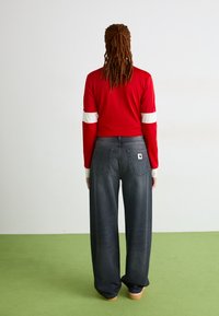 Top de manga larga roja con rayas blancas en las mangas, combinado con jeans oscuros de pierna ancha que tienen bolsillos traseros y un parche con el logo.