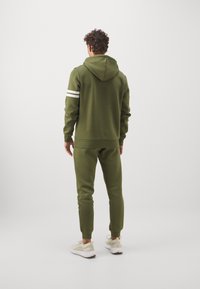 Olivegrüner Hoodie und Jogginghose aus weichem Stoff, mit weißen Streifen an den Ärmeln. Leichtes Design mit gerippten Bündchen und Taillenbund.