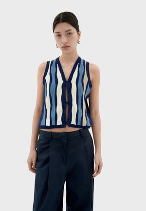 Knit vest featuring vertical blue, navy, and cream stripes, v-neckline, and ribbed edges, paired with high-waisted dark trousers.