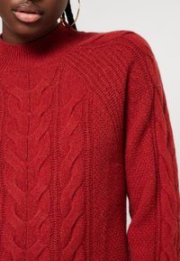 Gros plan sur une personne portant un pull en maille torsadée rouge et de grandes boucles d'oreilles en or contre un fond clair.