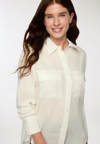Camicia bianca a maniche lunghe con bottoni, realizzata in un tessuto leggero. Presenta due tasche sul petto e un colletto classico. Tessuto liscio.