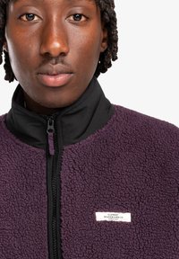 Fialová fleecová bunda s černým límcem, se zapínáním na zip a texturovanou tkaninou. Na přední straně se nachází štítek Element Skateboard Company.