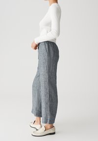 Femme portant un haut à manches longues blanc, un pantalon court gris ample et des mocassins blancs, se tenant contre un fond clair uni.