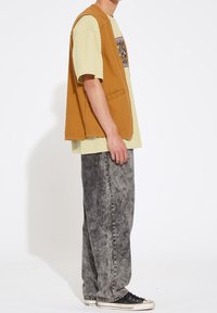 Modèle masculin portant une veste marron sans manches sur un t-shirt graphique jaune, un jean gris délavé à l'acide et des baskets noires basses, debout de profil.