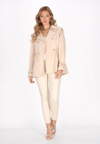 Trench coat beige chiaro con vita sfiancata, design doppiopetto, grande colletto e dettagli con bottoni. Abbinato a pantaloni color crema e sandali.