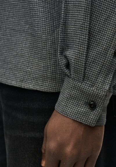 Κοντινό πλάνο ενός σκούρου χρώματος δέρματος χεριού που ξεκουράζεται δίπλα στο σώμα, φορώντας μανσέτα γκρι πουκάμισου με σχέδιο και μαύρο κουμπί.