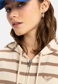 Sudadera con rayas beige y marrón, con cremallera frontal, tela texturizada, capucha ajustable y un logo de corazón en el pecho. Gorra azul llevada en la parte superior.
