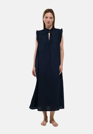 Femme debout pieds nus portant une robe maxi bleu marine sans manches, avec des volants aux épaules et un col clé noué.