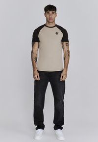 Beżowy dopasowany t-shirt z krótkimi czarnymi rękawami raglanowymi i logo na lewej piersi, w połączeniu z czarnymi dżinsami i białymi trampkami.