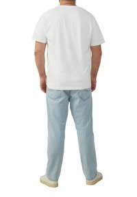 Man staat met de rug naar de camera, draagt een effen wit T-shirt met korte mouwen, lichtblauwe jeans en witte sneakers tegen een witte achtergrond.