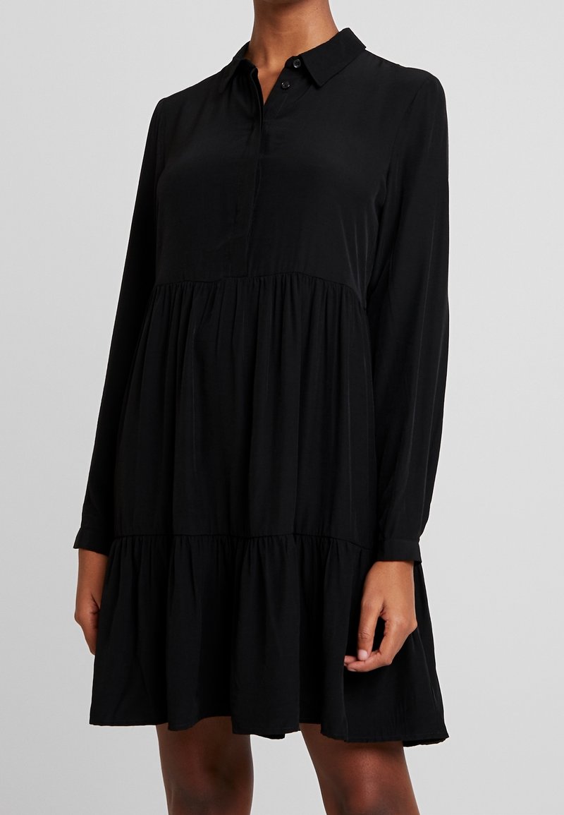 Robe noire à manches longues, col boutonné et bas à volants. Fabriquée en tissu doux à la texture lisse.