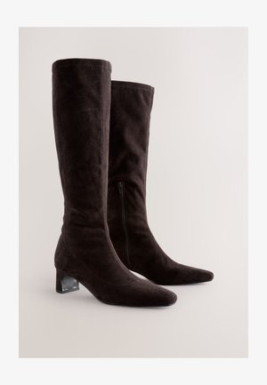 Bottes noires en daim montant jusqu'aux genoux, avec un bout pointu, un détail de fermeture éclair sur le côté et un talon bloc transparent. Texture lisse et silhouette ajustée.