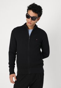 Svart stickad zip-up tröja med texturerat mönster, hög krage och ribbade manschetter. Bärs över en ljusblå skjorta.