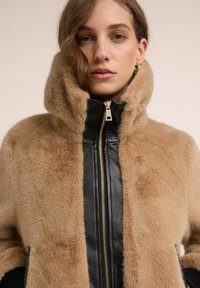 Kobieta w beżowej kurtce z sztucznego futra z wysokim kołnierzem i czarnym skórzanym suwakiem, patrząca prosto w kamerę.