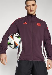 Burgunderfarget treningsjakke med røde detaljer, med hel glidelås og lommer på sidene, båret av en person som holder en flerfarget fotball.
