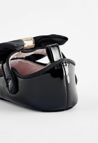 Schwarze Lackleder-Ballerinas mit einem weichen rosa Innenfutter, ausgestattet mit einer dekorativen schwarzen Schleife und einem goldenen Akzent nahe der Ferse.