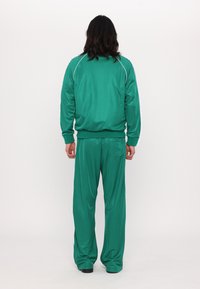 Tracksuit i levende grøn, lavet af glat materiale. Har hvide sidestriber på jakke og bukser. Løs pasform, elastiske manchetter og talje.