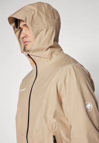 Beige huva jacka i lätt material, med en svart dragkedja och orange dragsko, samt en logo på vänster ärm.