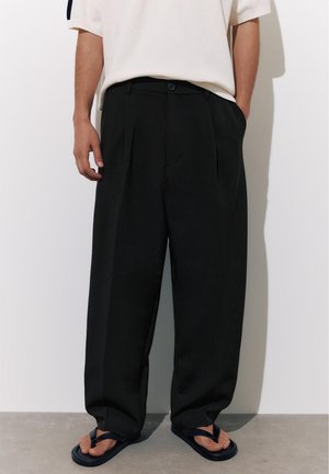 Osoba nosiaca voľné čierne plisované nohavice, biele tričko s krátkym rukávom a čierne žabky stojaca pri jednofarebnej stene.