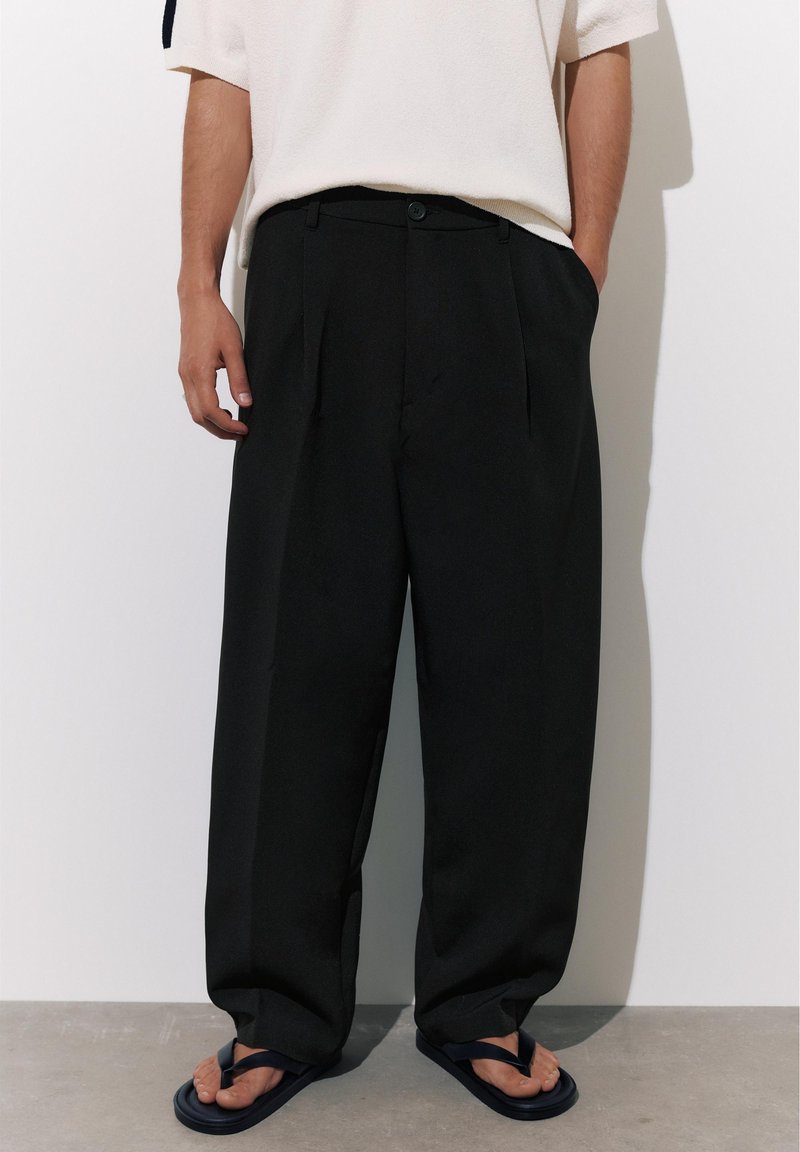 Personne portant un pantalon noir plissé ample, une chemise blanche à manches courtes et des tongs noires, debout contre un mur uni.