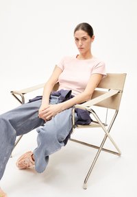 Lichtroze top met korte mouwen, denim wijde broek en lichtroze sneakers. Zittend op een beigevouwstoel met metalen frame.